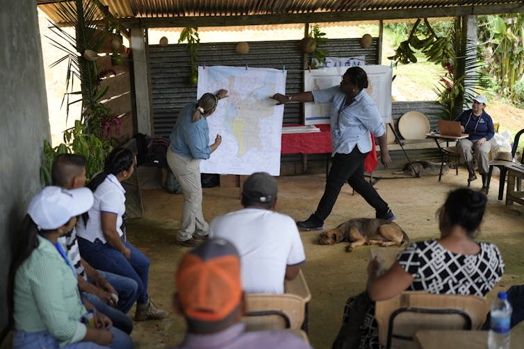 Two people point to maps during an outdoor community meeting.
