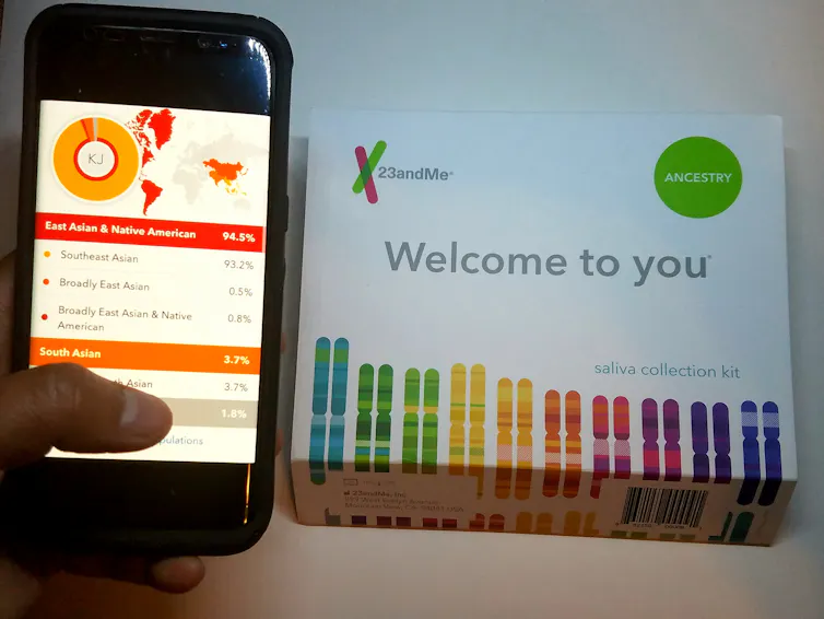 A smart phone displaying the results of a 23andMe ancestry test beside an unopened DNA testing kit