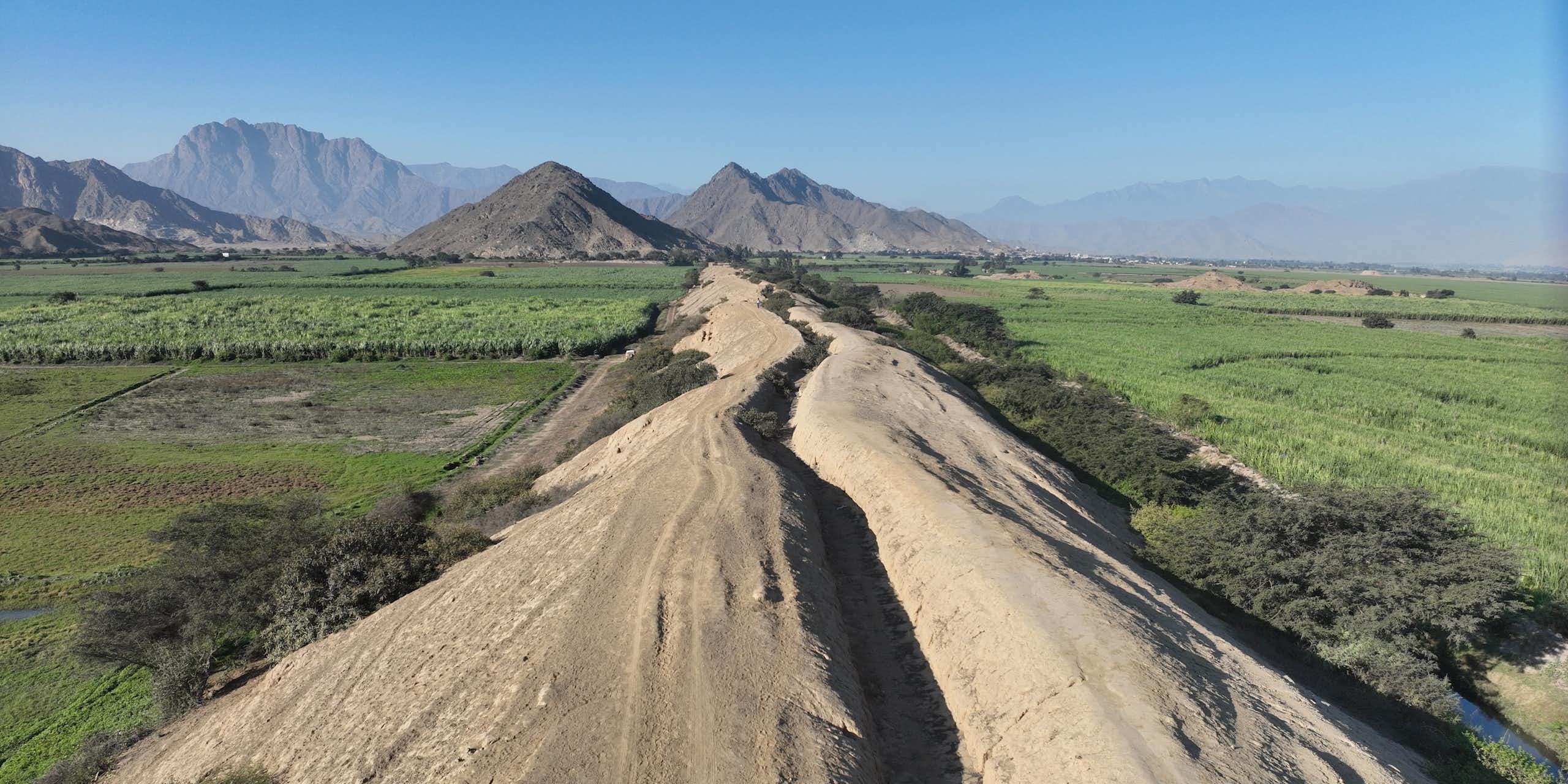 A dry canal through green fields with mountains in the distance