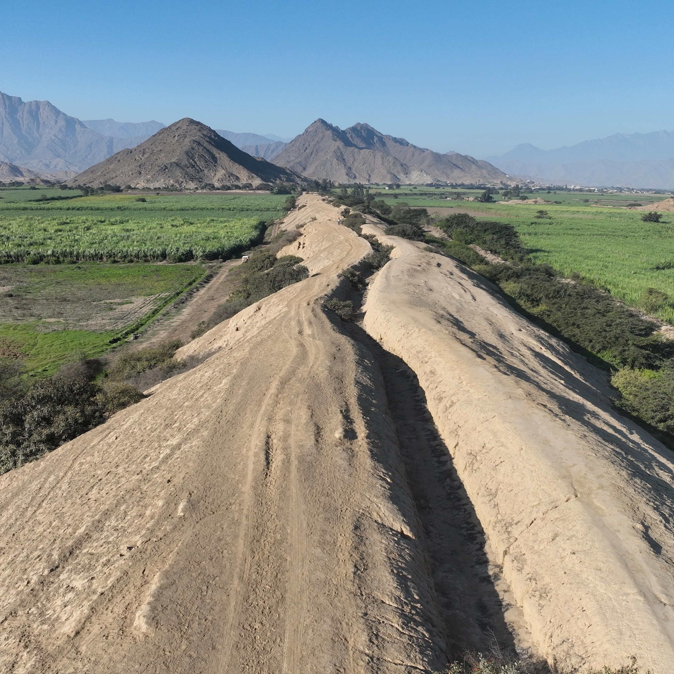 A dry canal through green fields with mountains in the distance