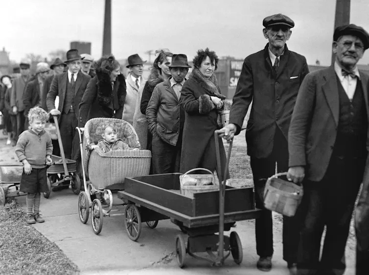 Black and white photo of people wearing clothing circa the 1930s standing in a line. Some stand with baby carriages, one has a wagon.
