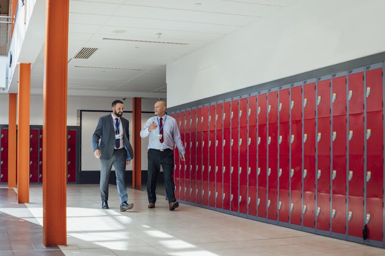Two men in suits walk past a row of lockers.