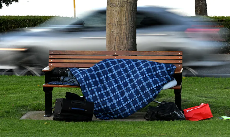 A person lying stretched across a park bench covered by a blue and white checkered blanket