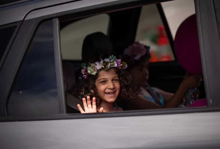 Una niña con flores en el cabello se encuentra en la parte posterior del auto y sonríe y ondas en la cámara