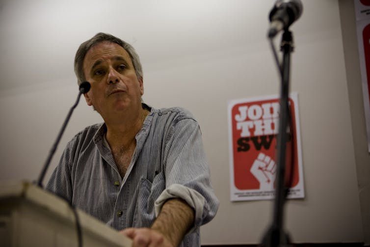 A man with shirtsleeves rolled up behind a podium, speaking into a microphone