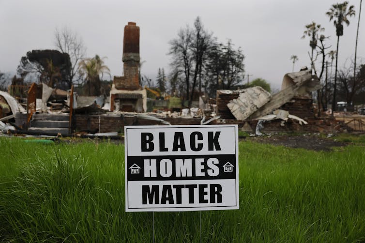 A sign reading 'Black Homes Matter' is posted in front of the ruins of a home