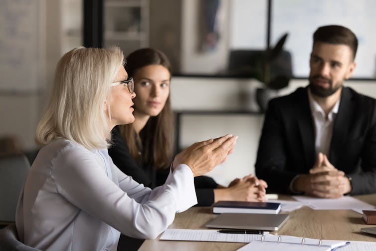 middle-aged woman at work speaking to younger colleagues around a desk.