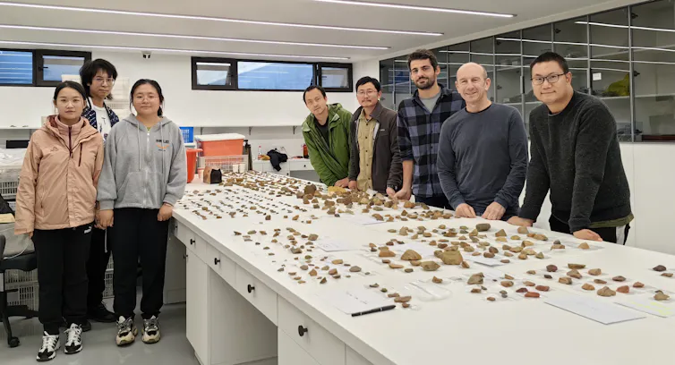 8 people stand beside high table displaying hundreds of small stone tools
