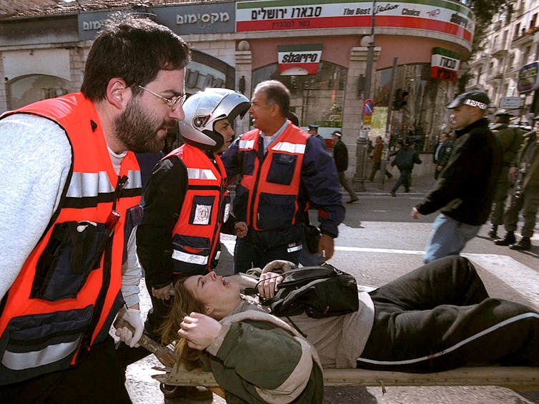 Men wearing orange vests carry a woman lying in pain on a stretcher.