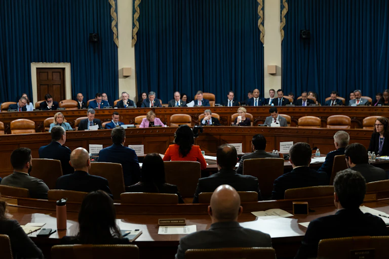 A crowd of people sit and face another group of people in a formal room with big brown chairs.