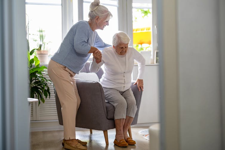 A middle aged woman helps an elderly woman get up from an armchair