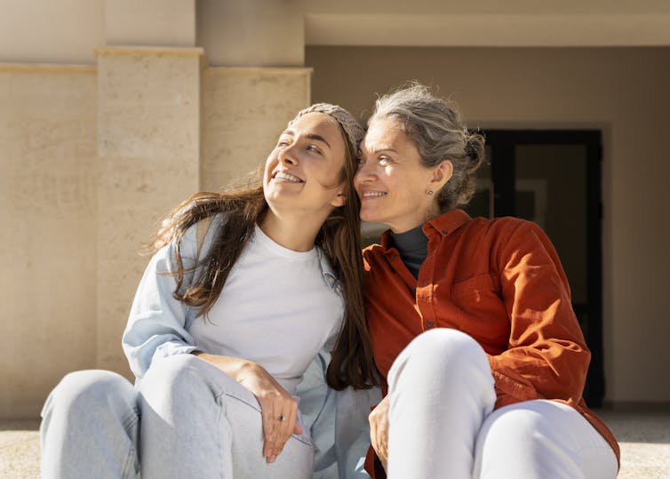 An older woman sitting outdoors with her arm around a younger woman, both smiling and looking at something in the distance