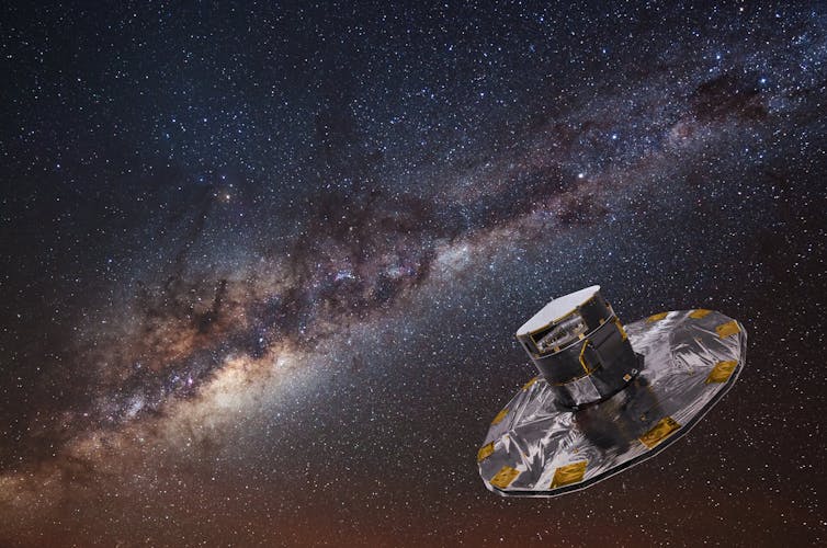 A background of stars on a dark sky including the beautiful, dusty stripe of the Milky Way. In the bottom left quadrant is the Gaia spacecraft. It looks like a top hat with a wide brim. It’s mainly silver in colour with gold patches.