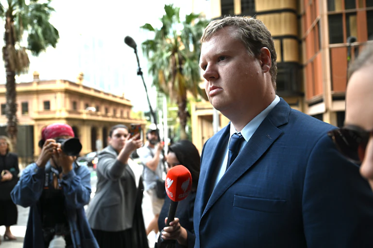 A man in a suit walks outside surrounded by a press pack