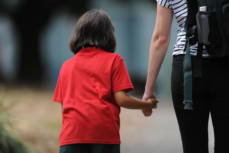 A woman holds the hand of a young child in school uniform.