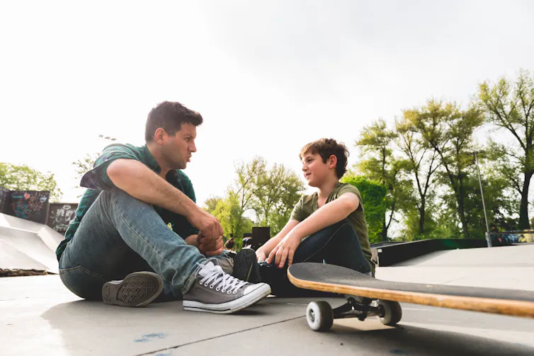 An adult man and child sit and talk at a skate park.