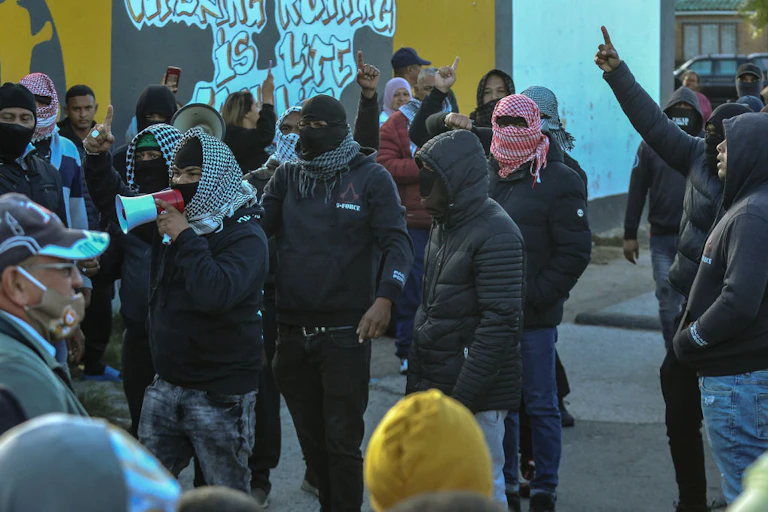 People with their faces obscured by scarves stand in front of a wall covered with graffiti, one of them speaking into a loudhailer