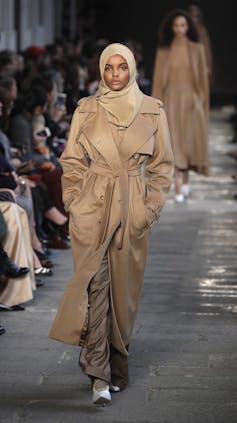 a female model in a beige trench coat and matching hijab walks the runway