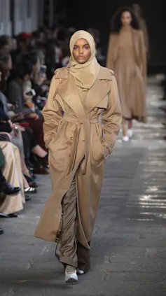 a female model in a beige trench coat and matching hijab walks the runway