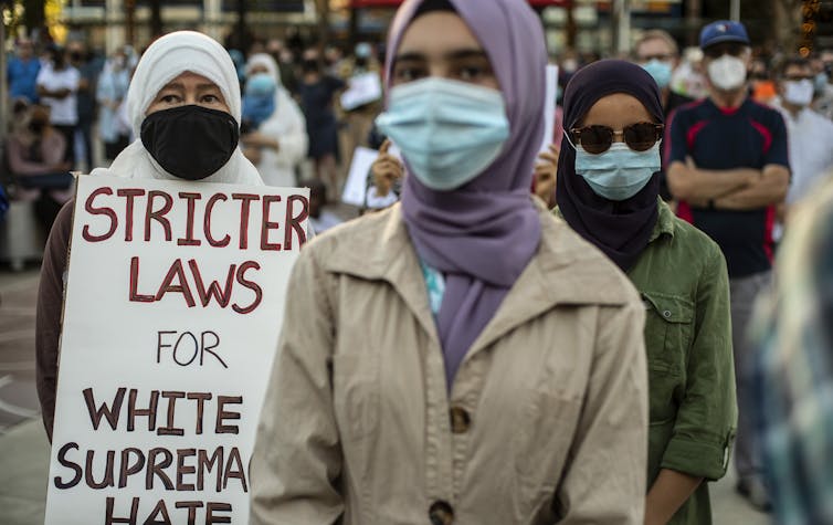 three woman at a protest stand in looking at the camera. one holds a sign that says 'stricter laws for white supremacist hate'