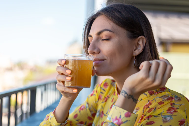 Headshot of woman sitting on a balcony, sniffing a glass of yellowy orange fluid
