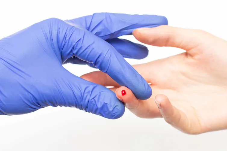A latex gloved hand holding a finger that has a drop of blood on it.