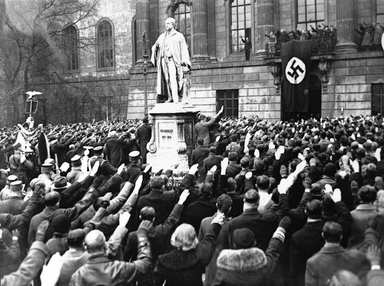 a crowd of people stand near a statue of a man holding their right hand up in a salute in front of a stone building