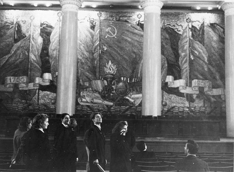 a group of students stand in a room with two big columns and a mural of soviet propaganda on the wall