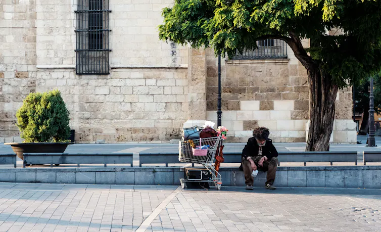 Unhoused man sits on the sidewalk with a grocery cart full of belongings.