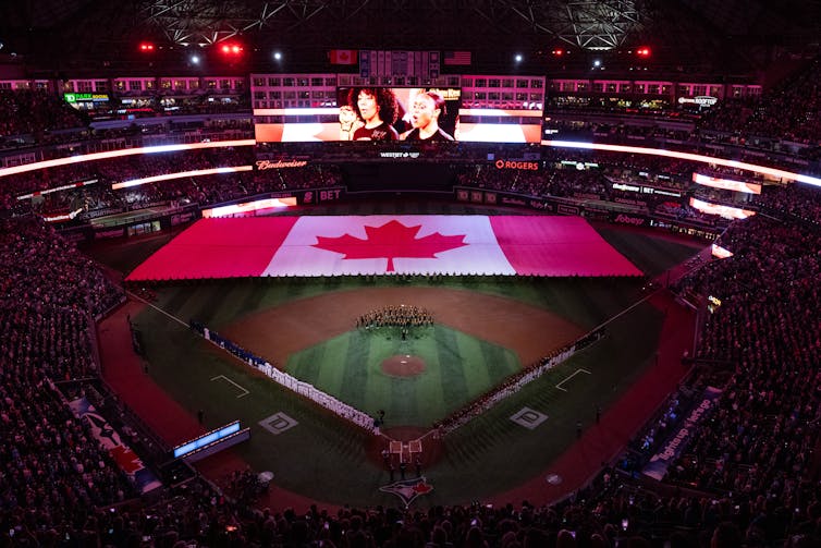 Una bandera canadiense masiva en un campo de bƩisbol oscuro, porque se ve a las personas cantando un himno en el Jumbotron.