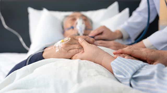 Photo d’un homme âgé intubé et allongé sur un lit, avec ses proches à côté de lui.