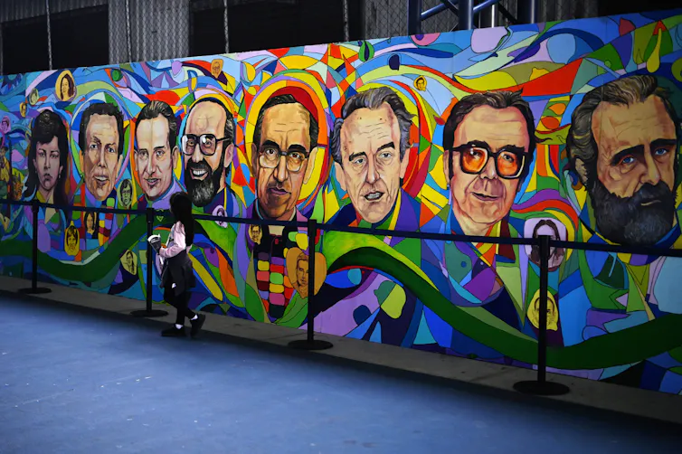 A girl stands in front of a multi-colored mural depicting the faces of eight people.