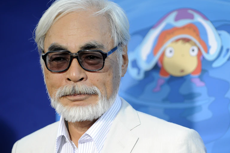 Man with white hair and tinted glasses, wearing a white suit and standing in front of an animated drawing.