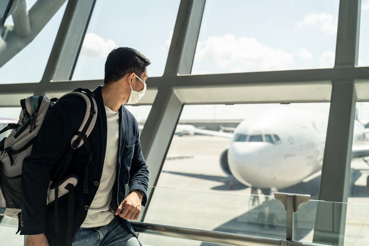 A man wearing a mask looking out an airport window at a plane.