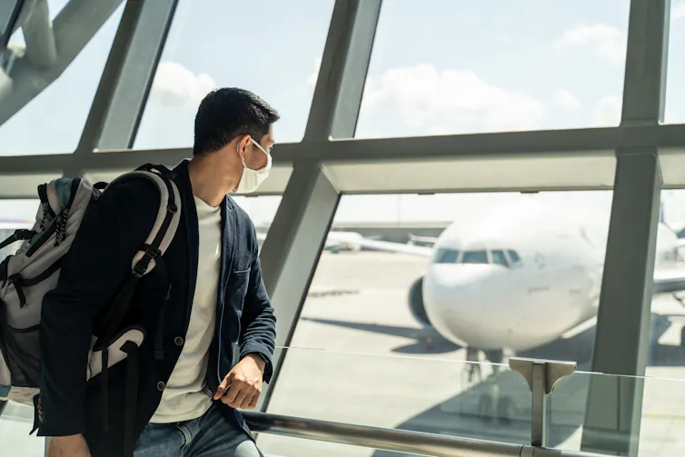 A man wearing a mask looking out an airport window at a plane.