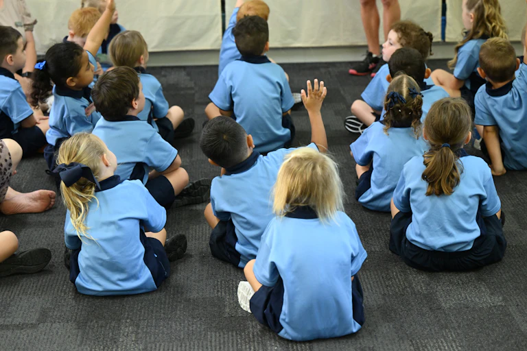 Young children sit in a classroom, one raises their hand.