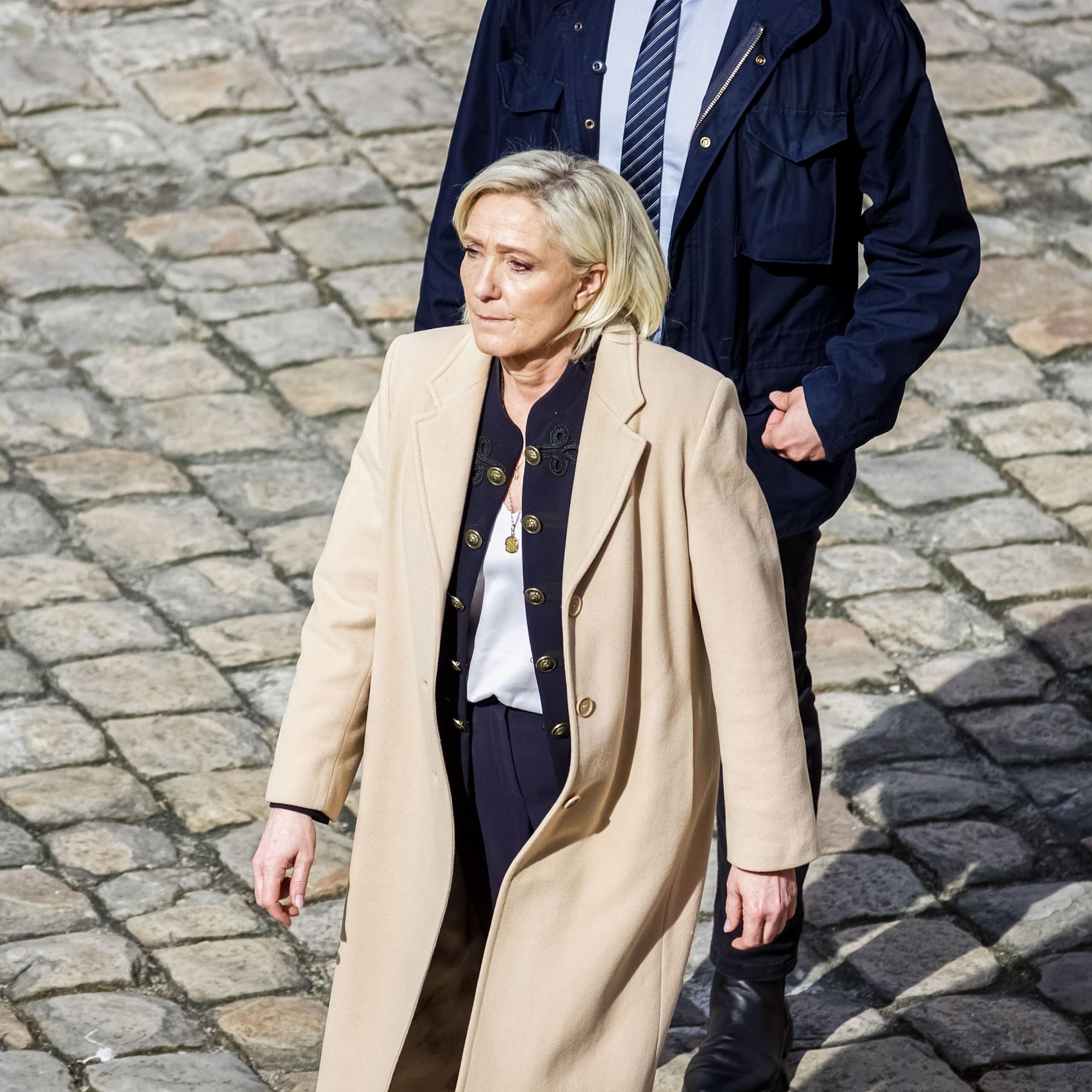 Marine Le Pen is walking on cobblestones with a man in a suit behind her.