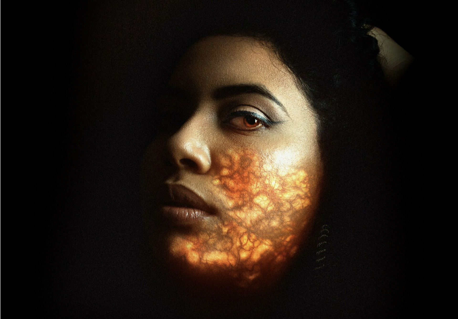 A close up of a woman's face in profile, eyeing the camera. On the lower part of her face is a projection of red and gold, like blood vessels.