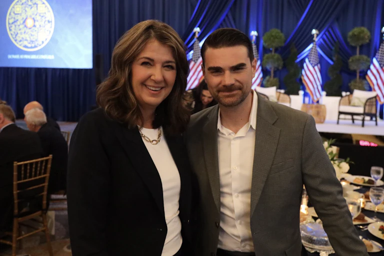 A dark-haired woman smiles standing next to a shorter man with dark hair and a beard.