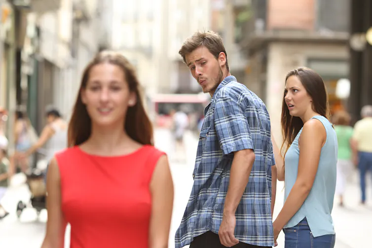 Young man ogles an attractive woman over his shoulder as his girlfriend looks annoyed.