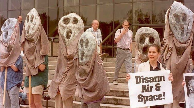 People hold large puppets of ghost-like figures and one holds a sign reading 'Disarm Air & Space!'