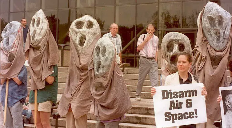 People hold large puppets of ghost-like figures and one holds a sign reading 'Disarm Air & Space!'