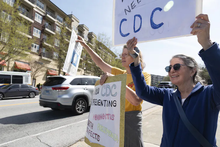 Two women hold protest signs about CDC layoffs along a roadside.