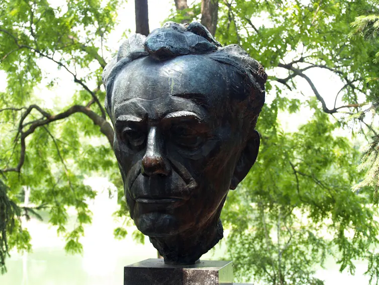 A large metal bust of a man's head, positioned against leafy green trees.