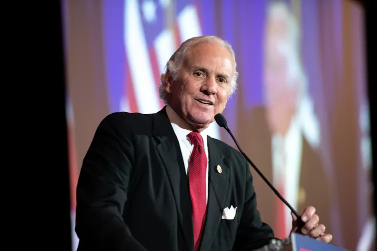 Man with gray hair in a suit and red tie speaks at a podium.