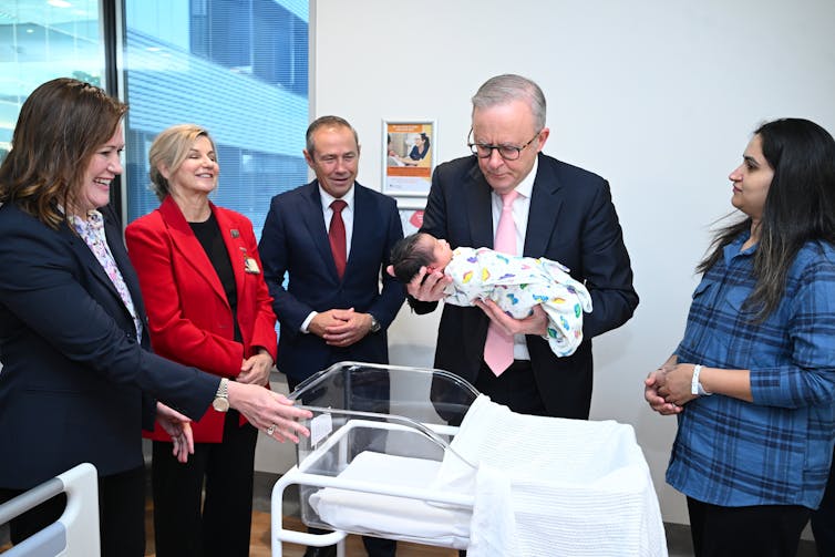Anthony Albanese holds a newborn baby above a hospital cot, surrounded by people