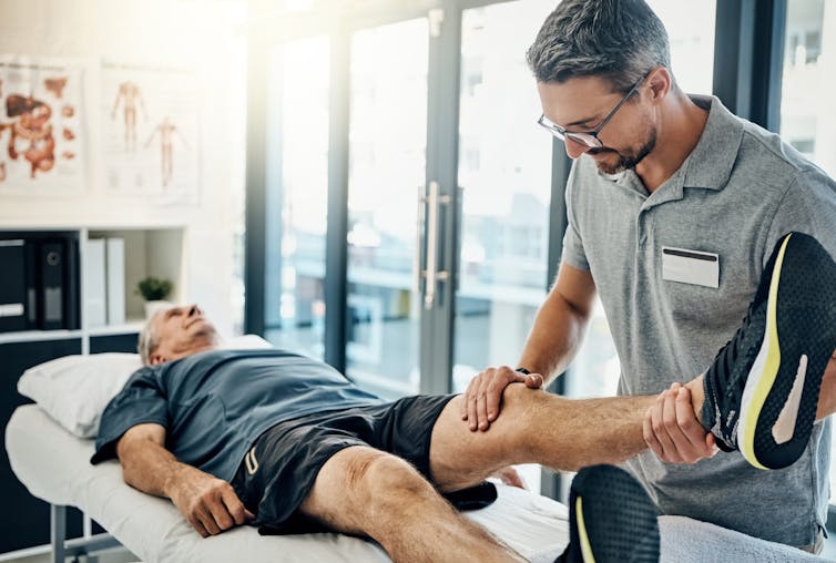 A senior man lying down while a physiotherapist examines his knee.