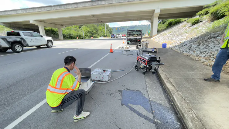 A researcher wearing a bright vest types on a computer that's connected to several pieces of equipment, on the shoulder of a road.