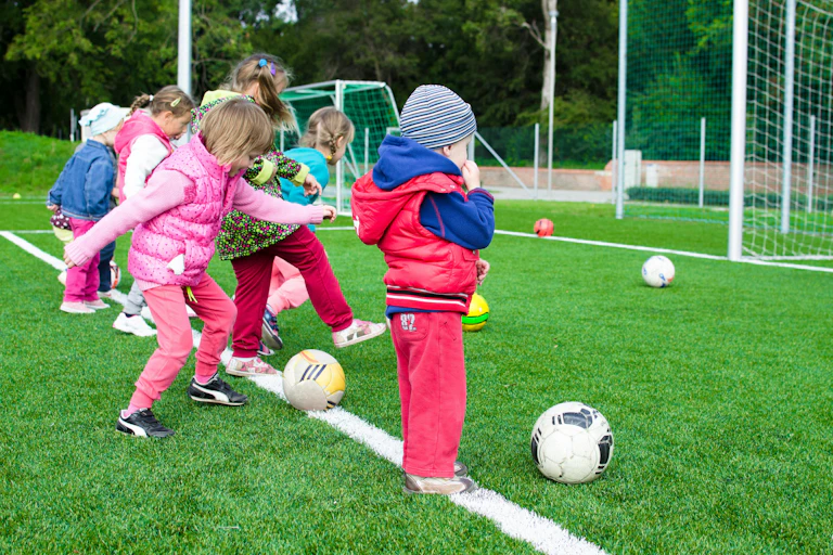 Young children stand in a line kicking balls on a field.