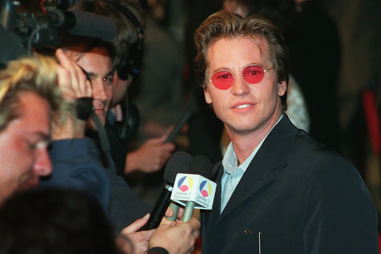 Actor Val Kilmer is photographed by paparazzi at a red carpet event in 1997. He has red shades on, and a mic in front of his face.
